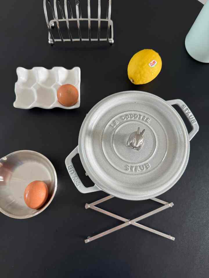 黒い背景にステンレスの折りたたみ鍋敷きとグレーのストウブ鍋、白い卵トレイと黄色いレモンが置かれたキッチン用品の写真