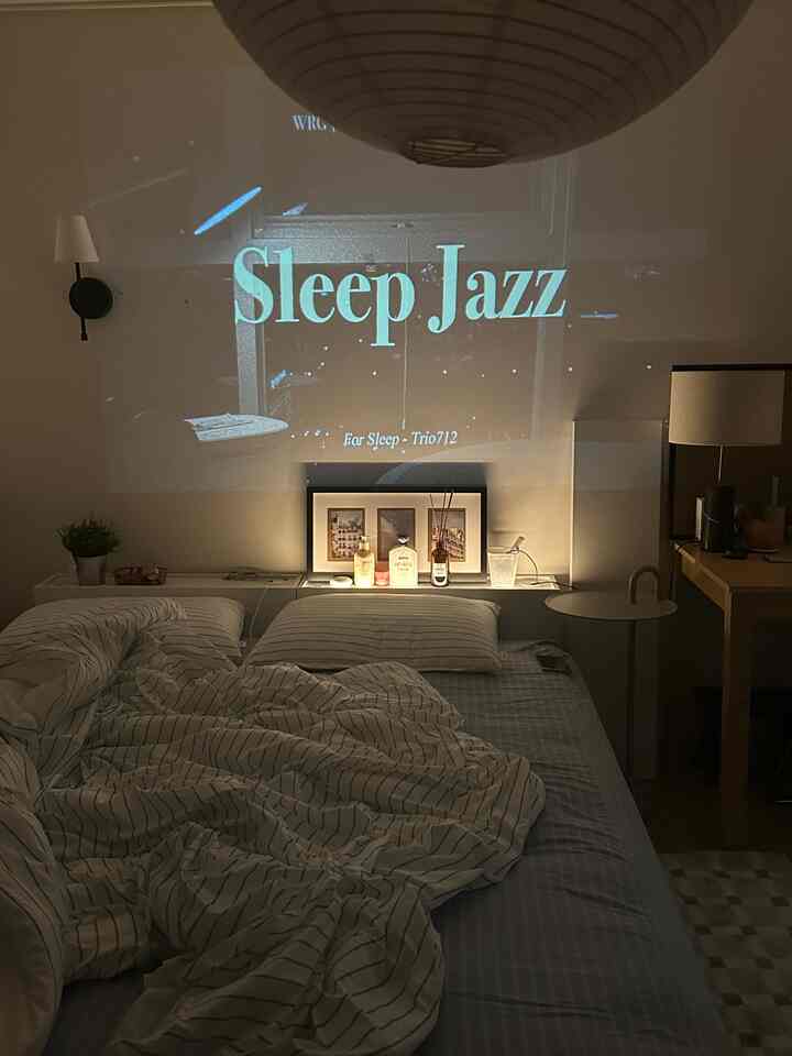 落ち着いたホワイトとベージュ調の寝室、ベッドとナイトテーブル、プロジェクターで壁に映された音楽映像のある雰囲気の良い空間