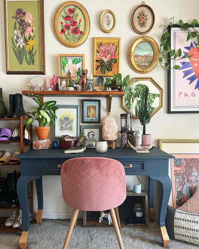 Vintage-inspired home office featuring a navy desk and pink velvet chair, adorned with numerous picture frames and lush plants for a cozy workspace