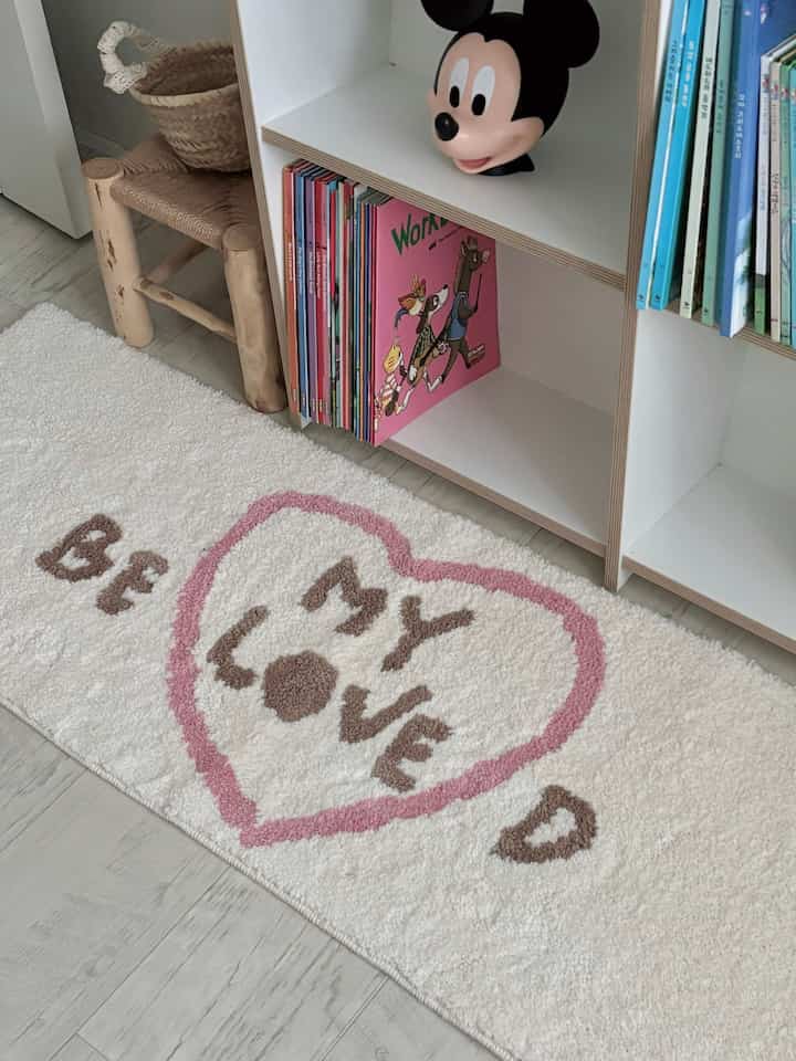 白とピンクを基調とした子供部屋、前面に本が並ぶ本棚と心温まる雰囲気のラグが配置されたインテリア