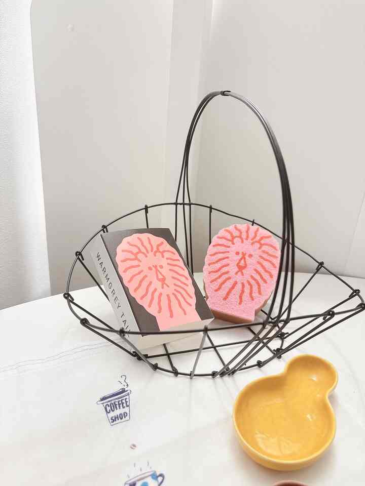 白い背景のキッチン空間に黒のワイヤーバスケット、ピンクのライオン型スポンジと黄色のミニプレートが配置された清潔感のあるインテリア