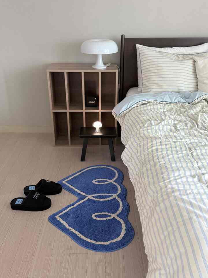 明るい木目調の床と白い壁の寝室、ストライプの寝具とシンプルなテーブルランプが配置された印象的な空間