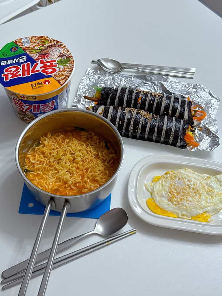 白いテーブルの上にラーメン鍋とキンパ、目玉焼き、カトラリーが置かれた質素な食事の様子