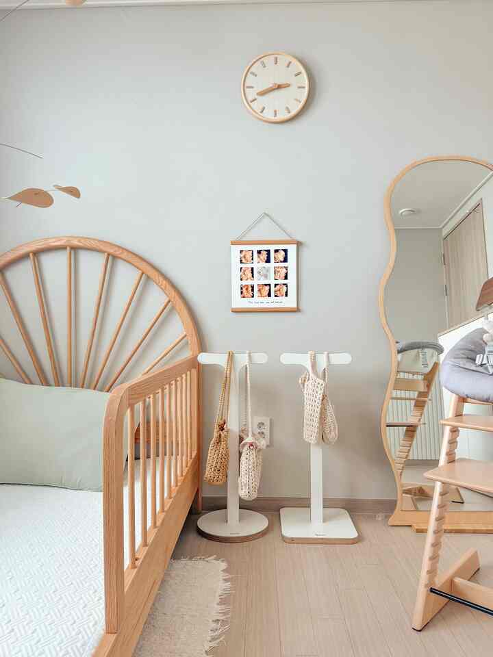 パステル調の壁と木目調の家具が調和した赤ちゃん用寝室、クリーム色の時計と波形の全身鏡が配置された温かみのある空間