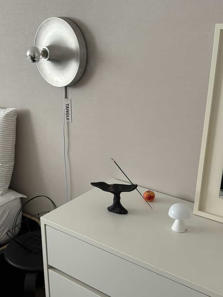 Clean bedroom with white and silver tones featuring a minimalist chest of drawers, decorative objects, and a wall sconce