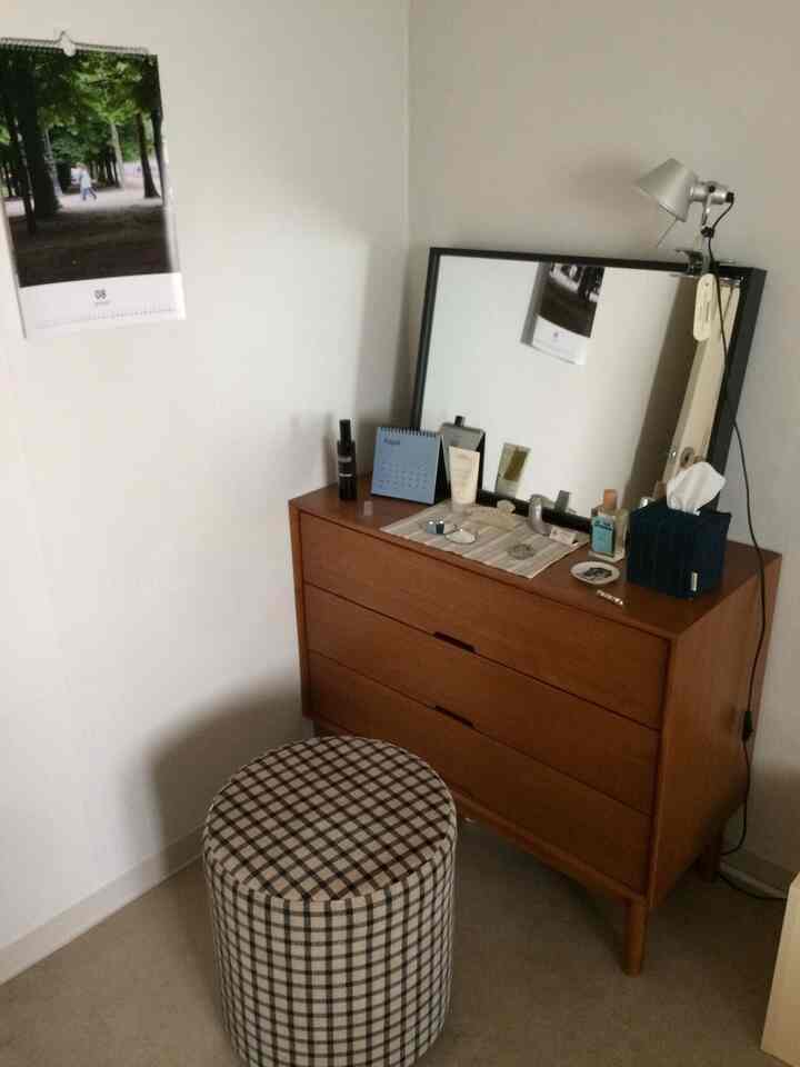 白い壁の狭い部屋にブラウンの化粧台と大きな鏡、チェック柄の丸スツールが配置されたシンプルな空間