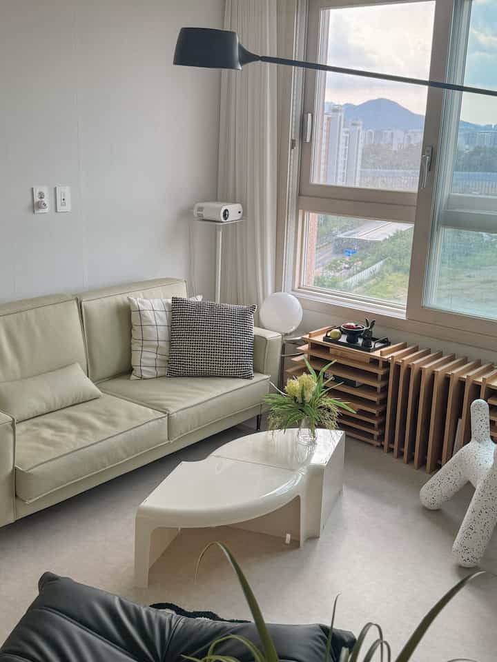 白とナチュラルカラーのリビング、ベージュのソファと白いコーヒーテーブル、窓際のプロジェクターが配置された印象的な空間