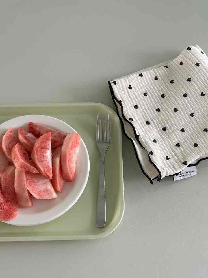 淡いグリーンのトレイに白い皿と桃のスライスがのせられ、右側には黒いハート柄のキッチンタオルが配置されたミニマルなキッチン小物の空間