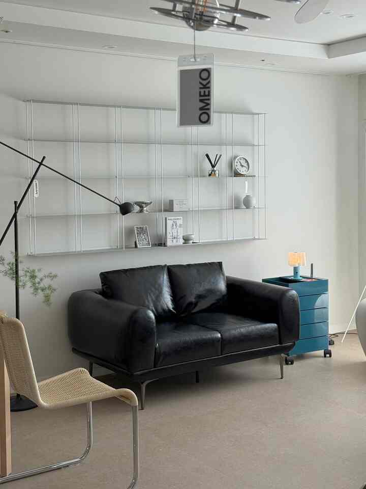 落ち着いたトーンのミニマルモダンなリビングルーム、中央にブラックレザーの2人掛けソファと金属製の棚、ブルーのサイドテーブルが配置された空間