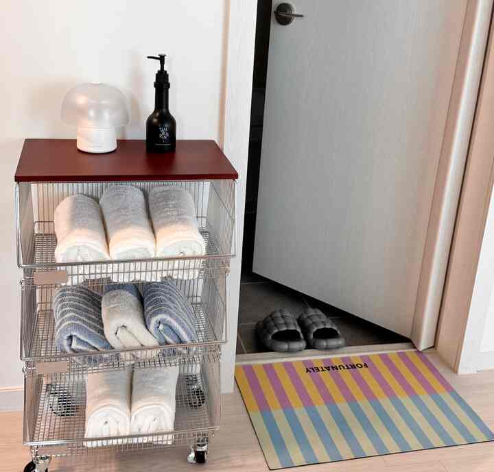 白と青のタオルが整然と並べられたメタル収納ワゴンと、開いたドアの隣にあるストライプの玄関マットが見える清潔な玄関