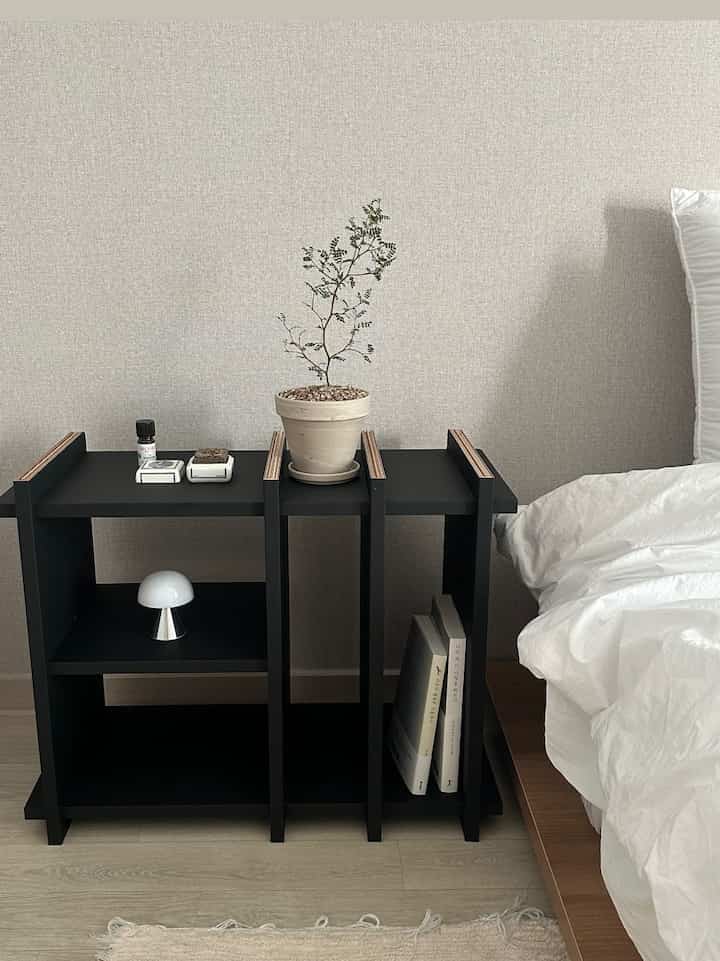 Natural-toned bedroom in white and black featuring a nightstand with plant and lamp in a minimal setting