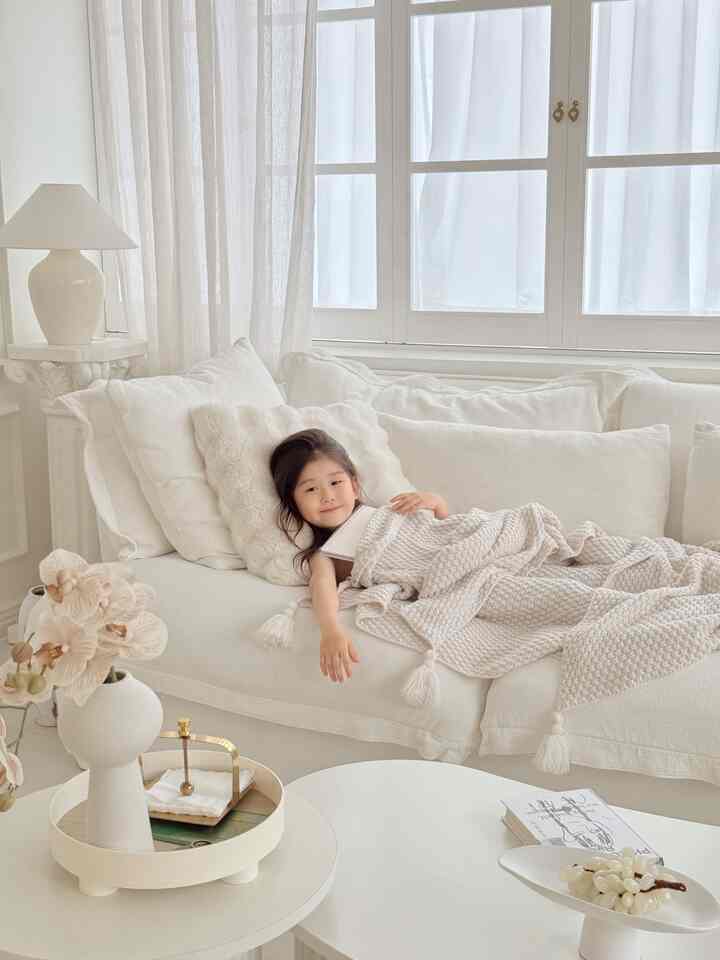 柔らかいアイボリー色調のリビング、白い大きなソファに子供が寝そべり、編みブランケットが配置された落ち着いた空間