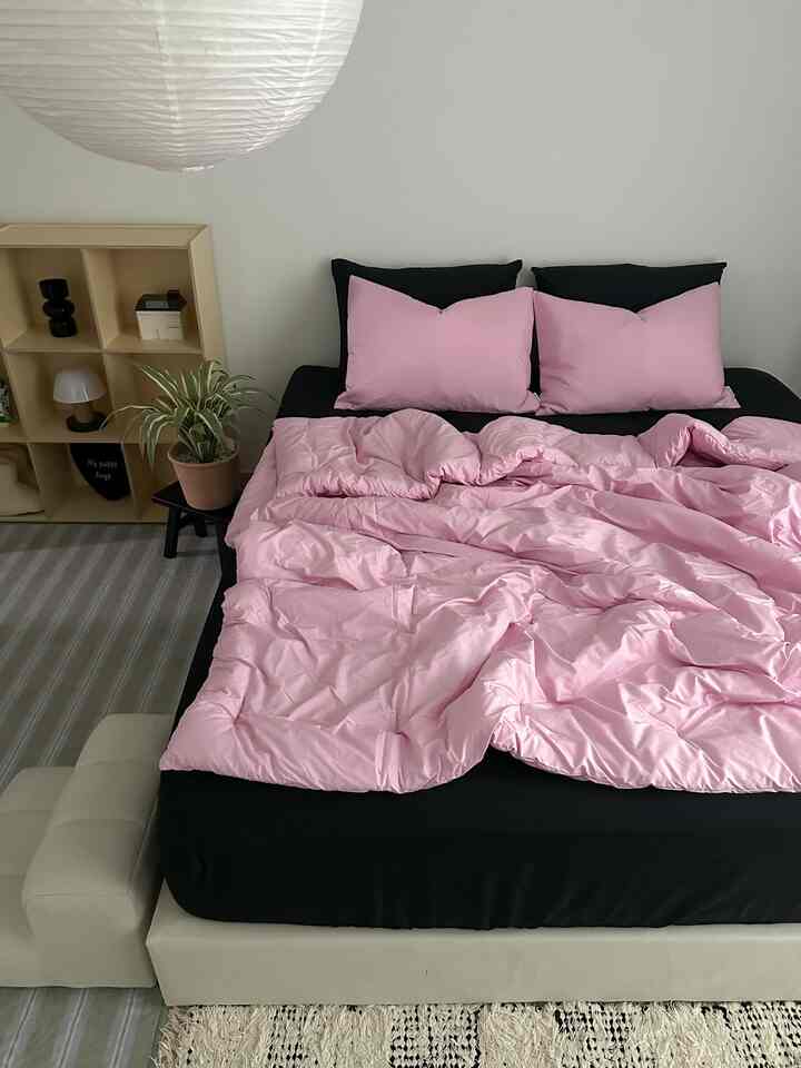 柔らかなピンクの寝具と黒いシーツが配置された寝室、ベージュの収納キャビネットと植物が調和した温かみのあるインテリア