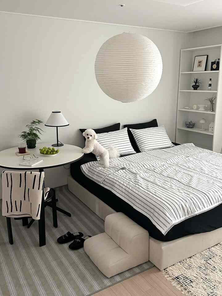 白と黒のトーンが調和した寝室、ストライプの寝具と丸テーブル、犬とペットステップが配置された清潔感のあるモダンな空間