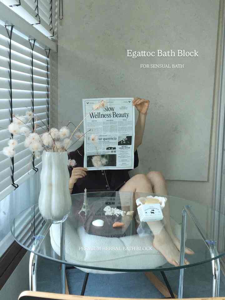 透明なガラステーブルと白いクッションチェアを配したモダンなバスルーム空間、読書する人とハーバルバスブロックが置かれている