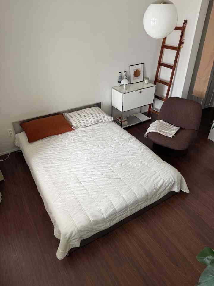 Warm modern bedroom featuring white walls, brown wooden floor, a bed with white bedding, and a brown lounge chair