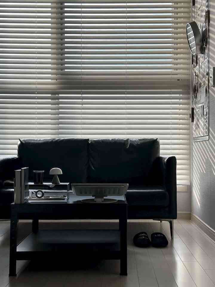 モダンな黒いソファとコーヒーテーブルが配置され、白いブラインドが印象的なリビングスペース