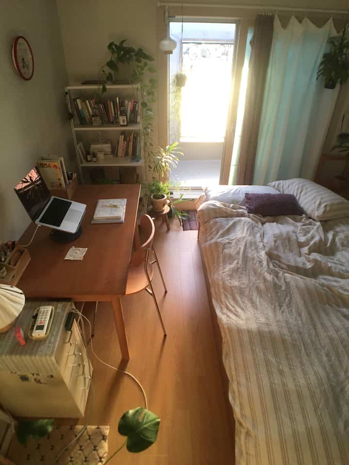 温かみのある木目調と自然光が差し込むワンルームの寝室、デスクと掛け布団カバーが居心地よく配置されたインテリア