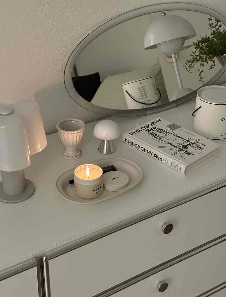 白を基調とした寝室のナイトテーブル、テーブルランプ、楕円形の鏡、灯されたキャンドル、本が配置された印象的な空間