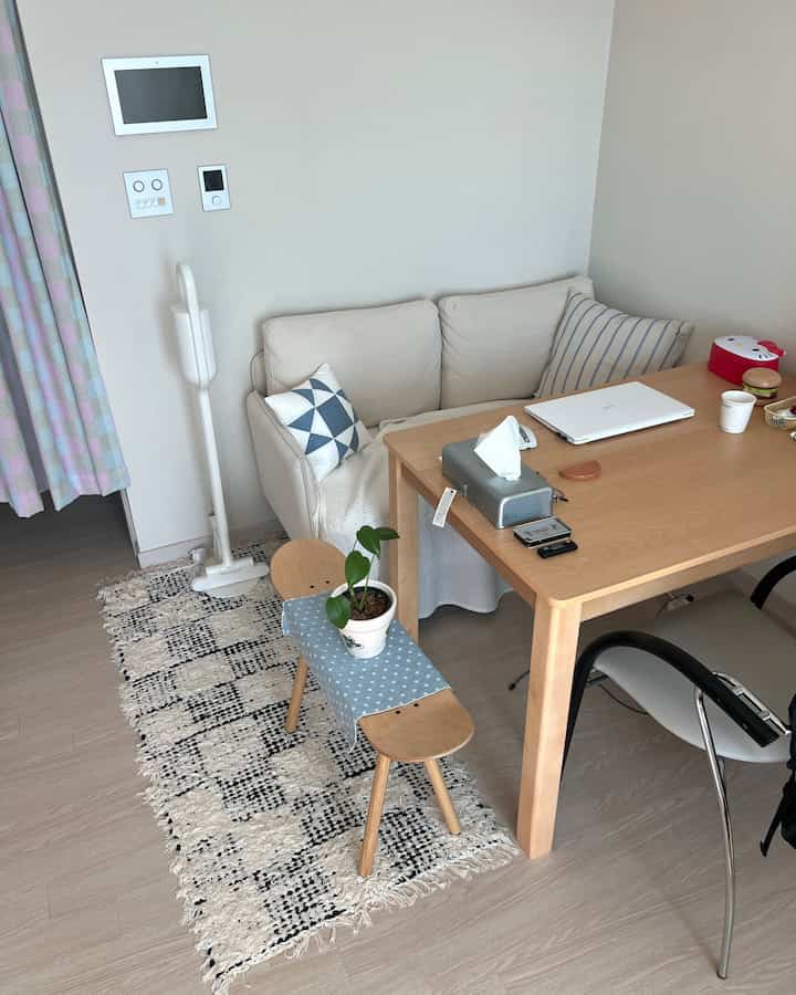 明るい白い壁とナチュラルな木製ダイニングテーブル、ソファとスツールが配置された一人暮らし向けの居心地の良いリビングルーム