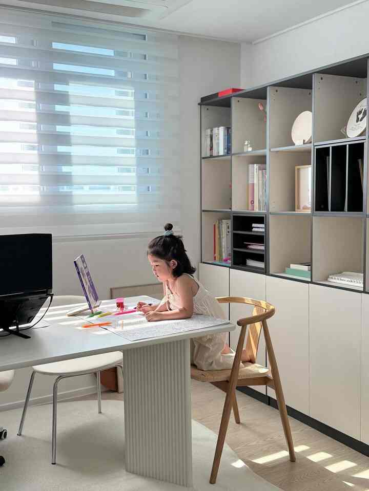 白と木目調が調和した書斎で、子供が机に座り読書や塗り絵を楽しむミニマルで自然な雰囲気のインテリア