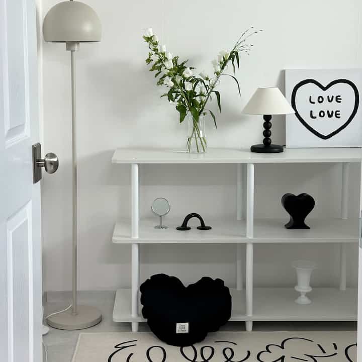 モノトーンの小さな部屋、白い棚とフロアランプ、ハート形クッションと花瓶が配置されたシンプルモダンな空間