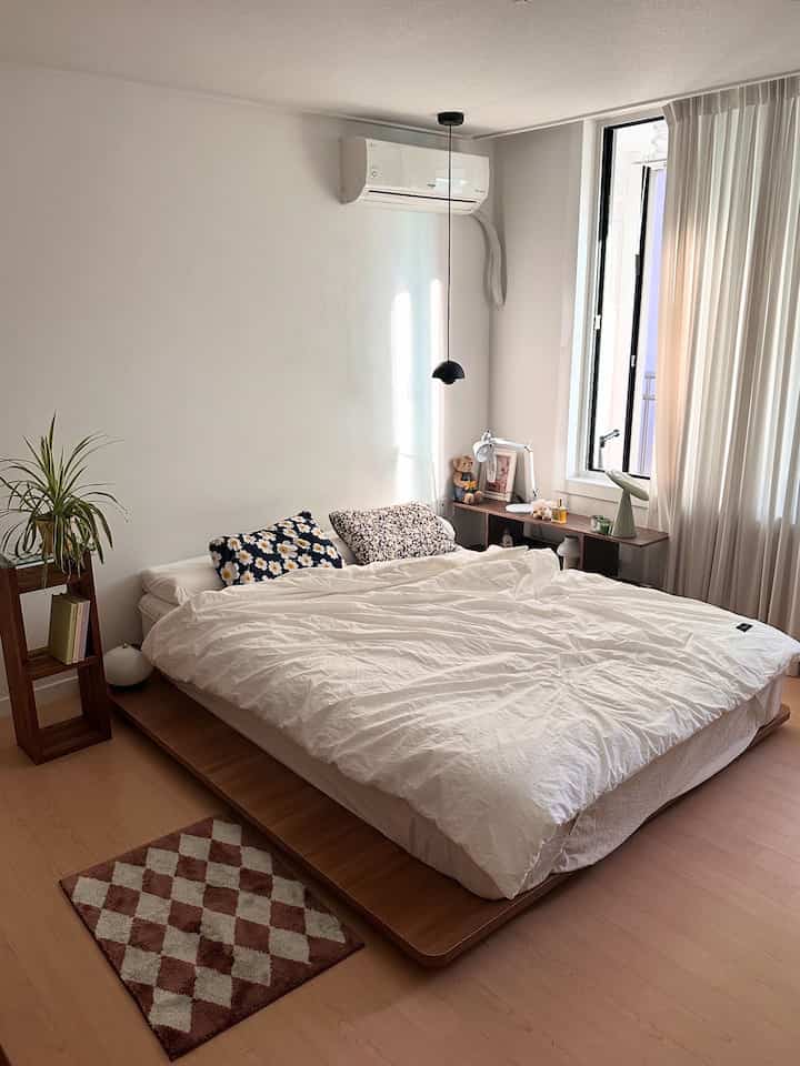 A bedroom dominated by white and wood tones featuring a low-profile bed, plant, and pendant light in a simple, natural setting