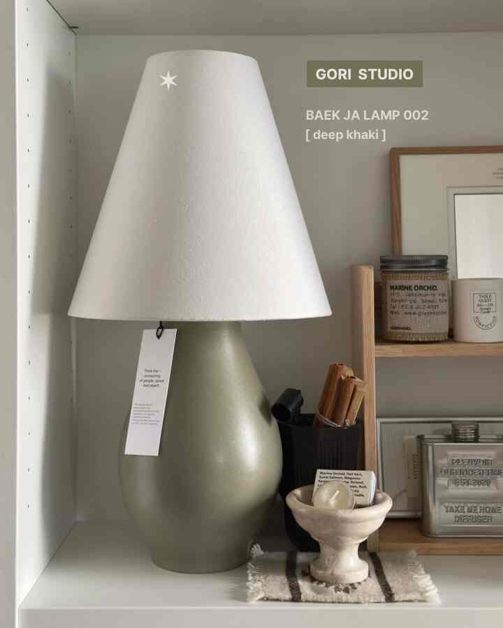 白色のランプシェードとカーキ色のボディが調和するテーブルランプが配置された棚のあるインテリア