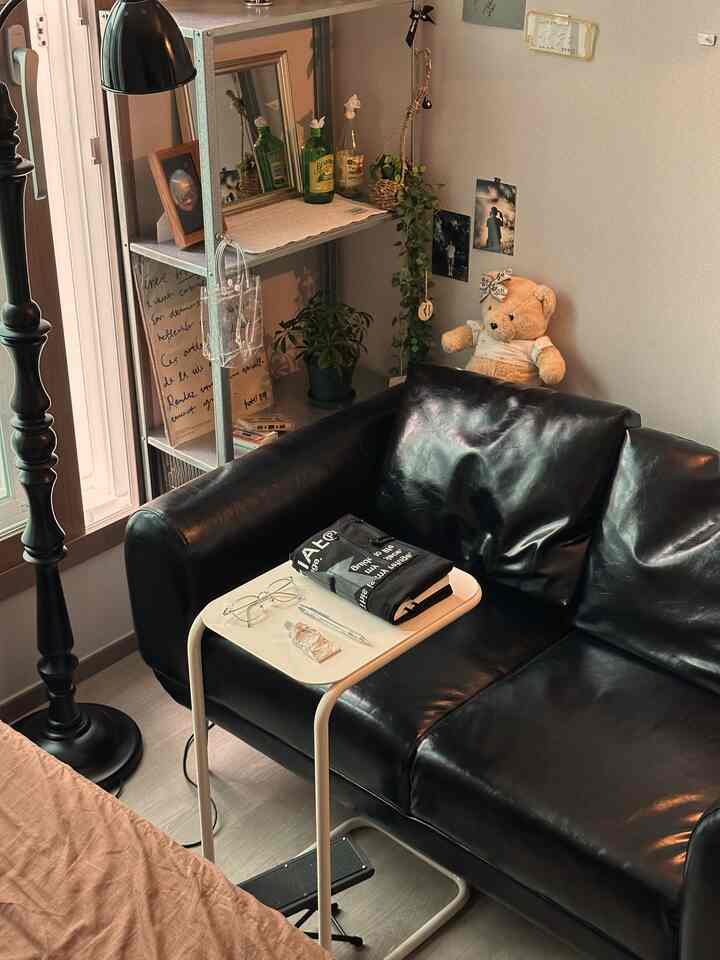 黒いレザーソファと小さな白いテーブルがあるコンパクトなワンルームリビング、植物と鏡で飾られた落ち着いた空間