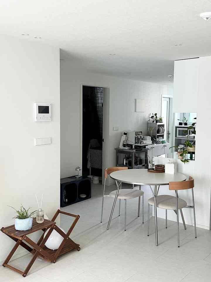 ホワイトとシルバーのトーンのリビング兼ダイニング空間、ラウンドのシルバーフレームのダイニングテーブルと木目調の背もたれ椅子が配されたミニマルなインテリア