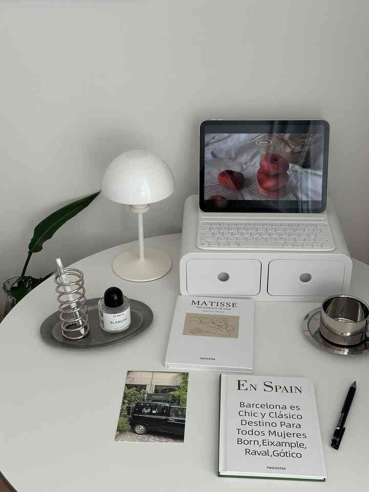 白を基調としたミニマルな小さなホームオフィス、丸い白いデスクにテーブルランプ、タブレット、本が整然と配置された雰囲気のあるインテリア
