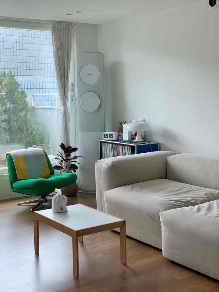 白い壁と木目調の床が特徴のナチュラルなリビング、クリームのソファと緑のアームチェア、中央に白いテーブルが配置された雰囲気のあるインテリア