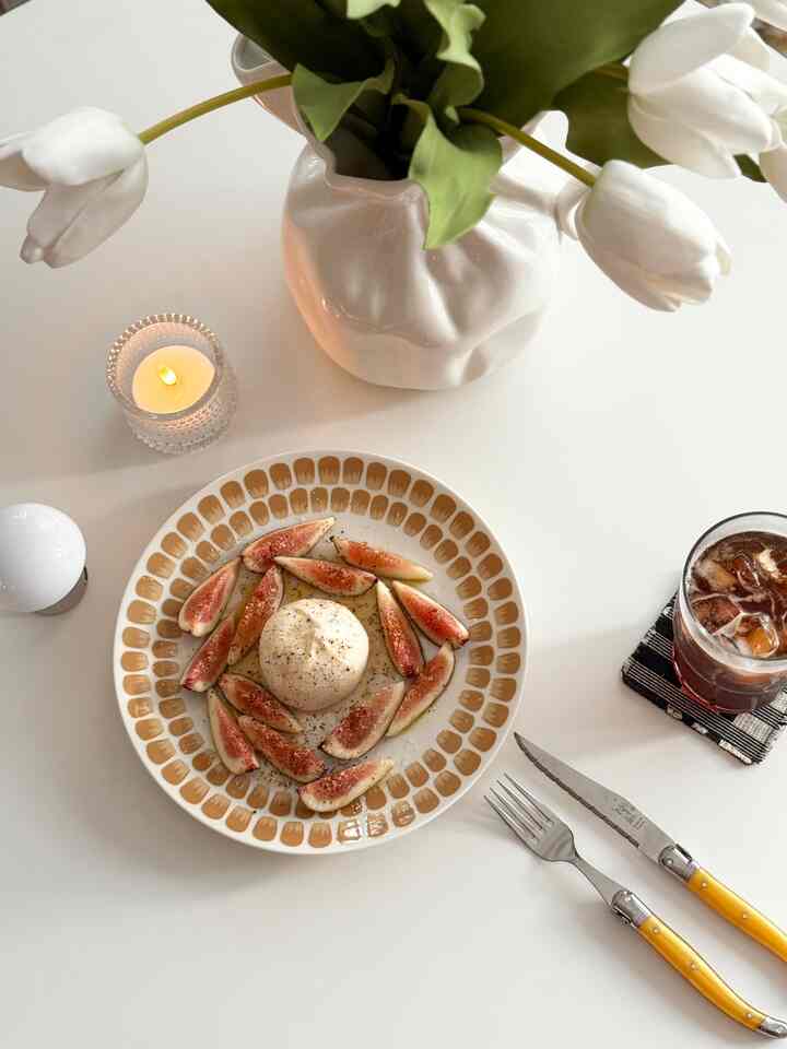 White table featuring a beige patterned plate with fruit, mood candle light, and white tulip vase in a simple home cafe setting