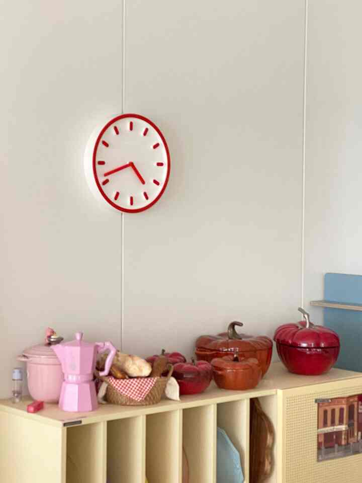 Natural tone kitchen with a white wall featuring a red-framed clock, cream-colored storage with red and orange pumpkin-shaped cookware displayed on top