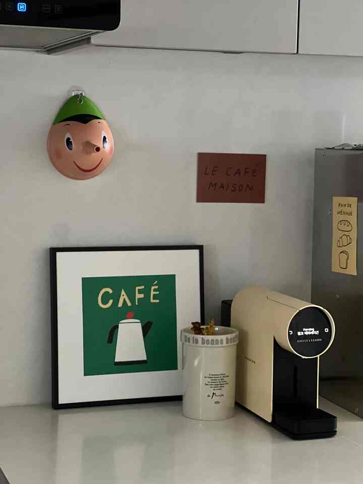 ホワイトの壁とベージュのミニカプセルコーヒーマシンがあるキッチンコーナー、ポスターと装飾品が温かみのあるおうちカフェの雰囲気を演出