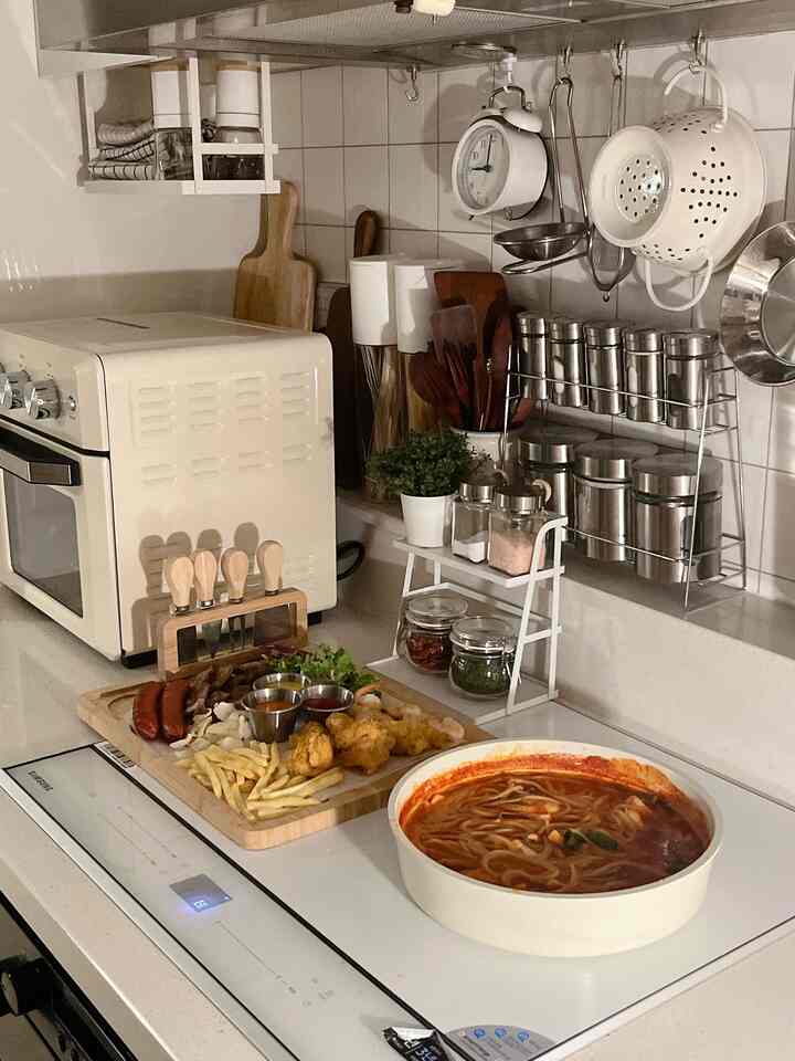 白とベージュの色調のキッチン、エアフライヤーと調理器具が整頓され、チーズプラッターと辛いパスタが皿に盛られた印象的な空間