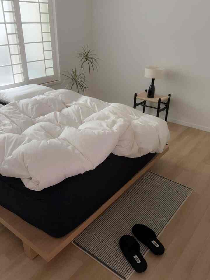 ホワイトとウッドトーンが調和したシンプルな寝室、白い布団と木製ベッドフレーム、小さなテーブルランプと植物が配置された雰囲気のあるインテリア