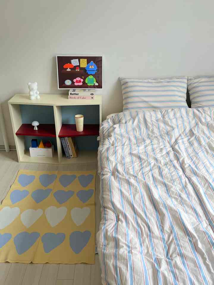 Natural and white toned 6畳 bedroom featuring a bed, bookshelf, poster, and rug arranged in a simple, baby-friendly setting