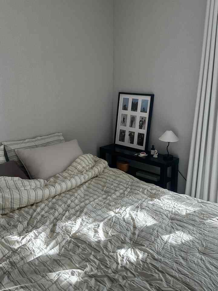 グレーの壁と白いカーテンがあるシンプルな寝室、黒いナイトスタンドとパターンの寝具が配置された落ち着いた空間