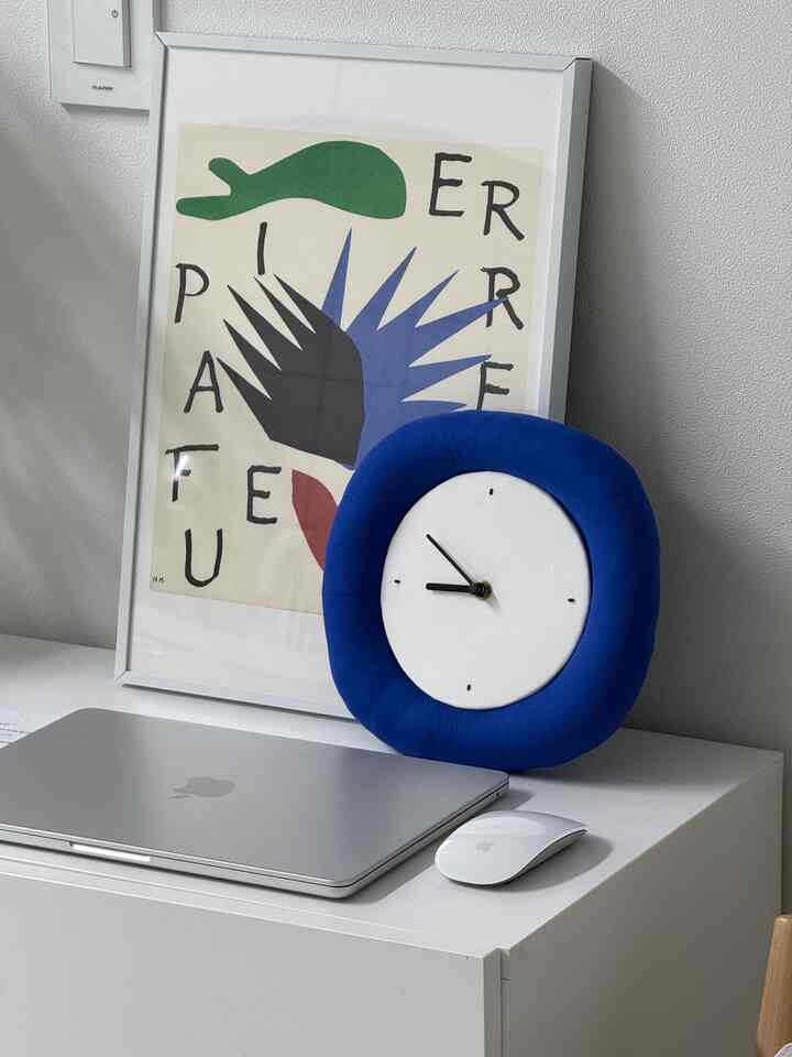 ホワイトトーンの壁とデスクにシルバーのMacBook Proと鮮やかなブルーの額縁時計が配置されたミニマルなホームオフィス