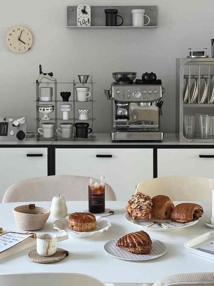 グレーと白の落ち着いたトーンのモダンなキッチン、中央にステンレス製のコーヒーマシンと多彩な食器が配置された雰囲気のあるインテリア