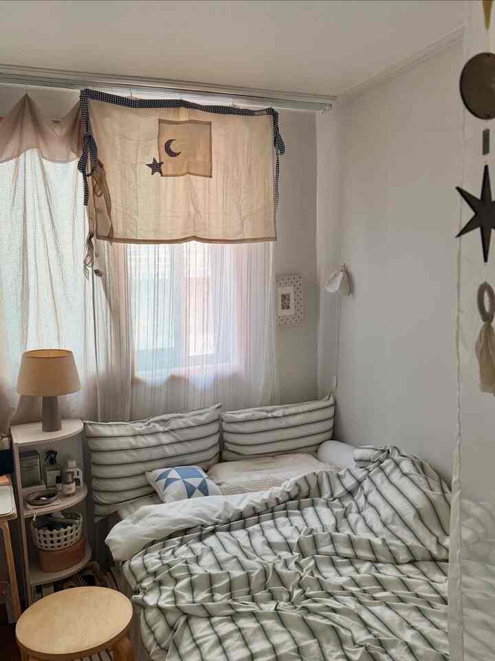 ホワイトとナチュラルトーンの寝室、星と月のパッチワークカーテンとストライプの寝具が配置された雰囲気のあるヴィンテージインテリア