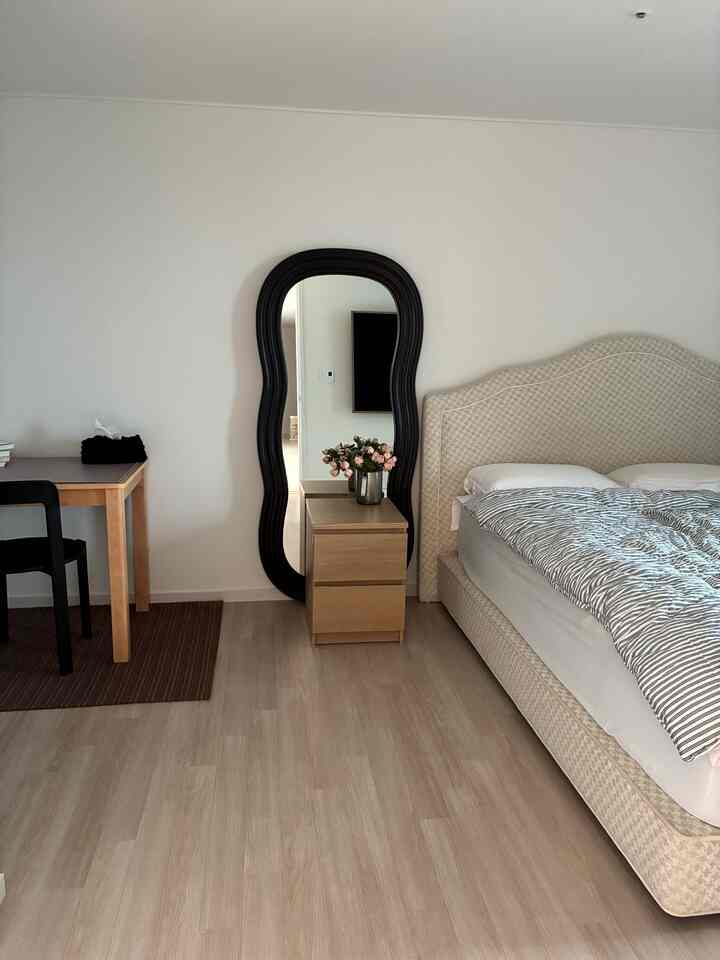 ホワイトの壁とブラウンの床のナチュラルな寝室、ベッドのストライプ柄寝具、黒縁の全身鏡、木製ナイトテーブルとデスクが配されたシンプルな空間