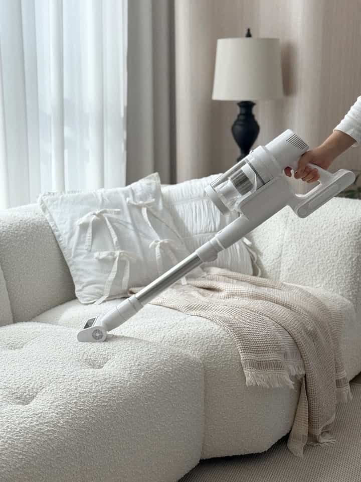 明るいホワイトのトーンのリビング、ファブリックソファの上をコードレス掃除機で掃除している様子と遮光カーテンが配置された空間