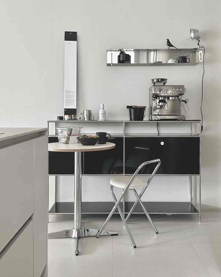 A simple modern kitchen and dining area in white and black tones featuring a stool, small table, and a home cafe setup with an espresso machine