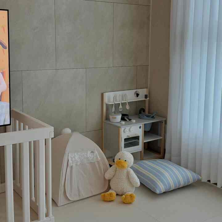 ベージュ色調のリビングの一角、ベビーベッドと子供用キッチン遊びセット、ペットハウスのテント、白いカーテンが配置された雰囲気のあるインテリア