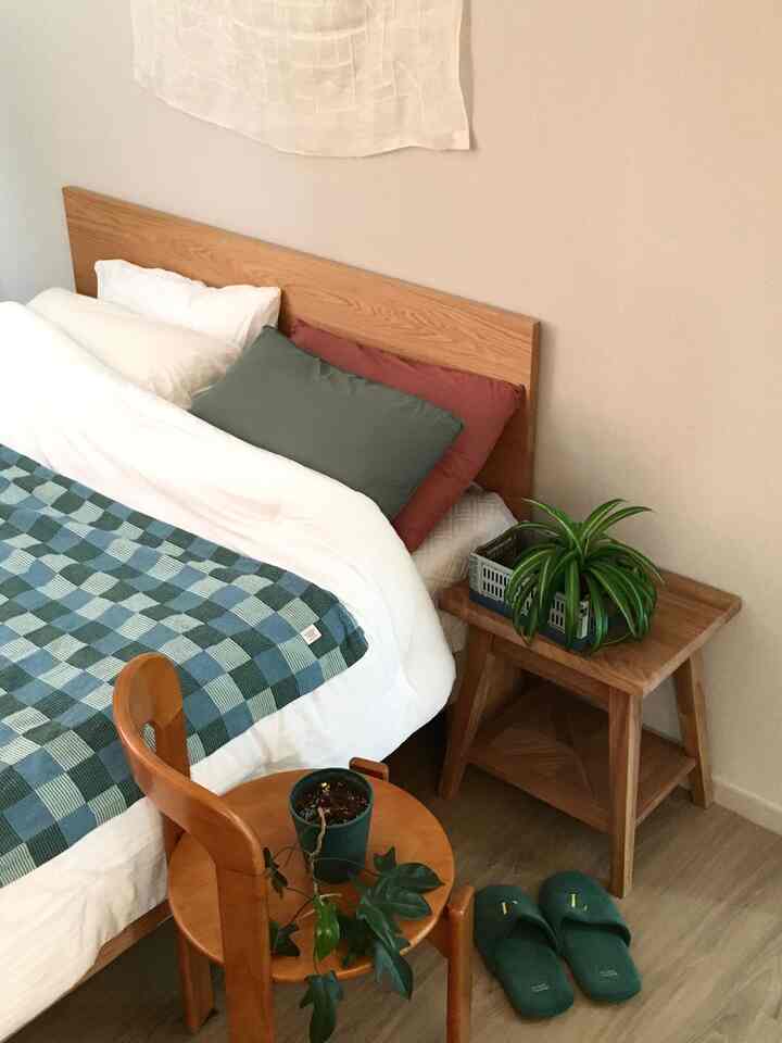 木目調の家具と植物、ナイトテーブルが配されたナチュラルな寝室空間、ベッドには白とチェック柄の寝具があり、落ち着いた雰囲気のインテリア