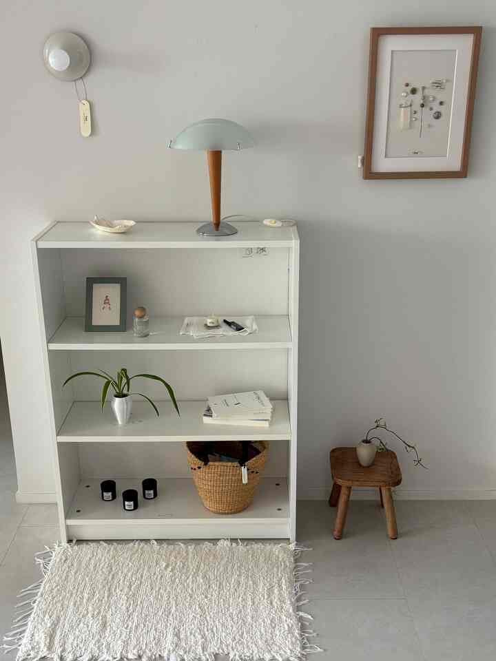 ホワイトとナチュラルカラーの小さな空間、白い本棚とラグ、木製スツールと照明が調和するミニマルなインテリア