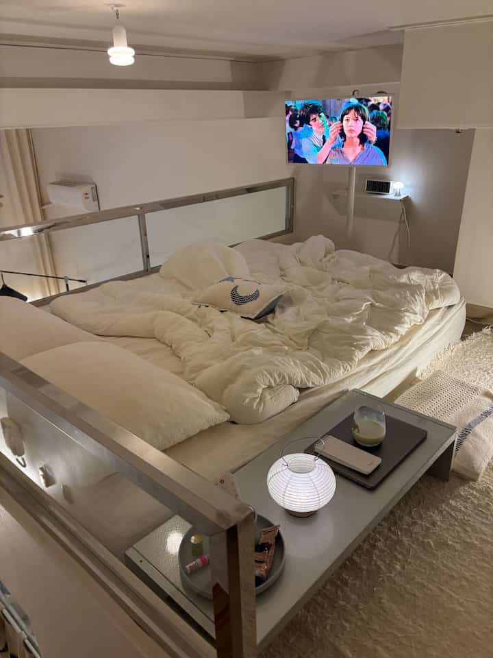 アイボリーとホワイト色調のロフト寝室、中央に大きなベッドとムードランプ、壁掛けテレビが配置された雰囲気のある空間