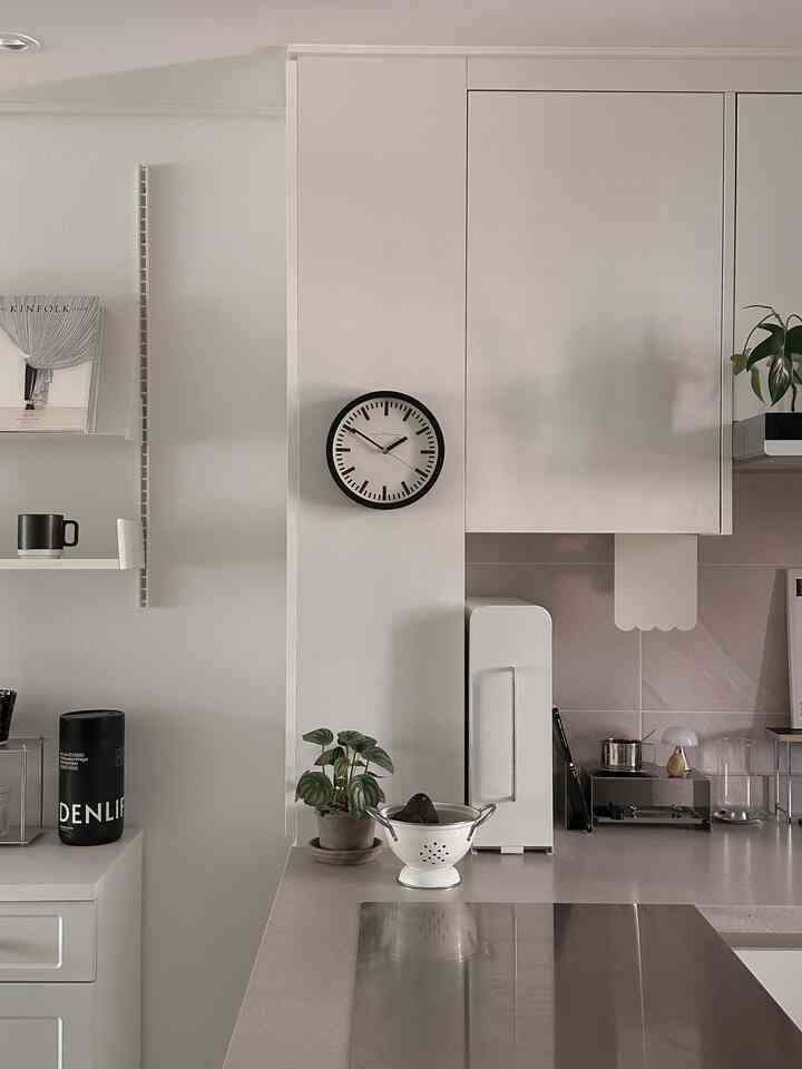 白と黒の調和が取れたキッチン、中央の壁に時計と植物、まな板が配置された清潔感のある空間